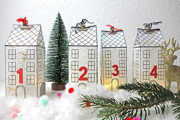 Four embroidered Advent houses numbered 1 to 4, lit from inside, next to a small bottle-brush tree and a golden deer