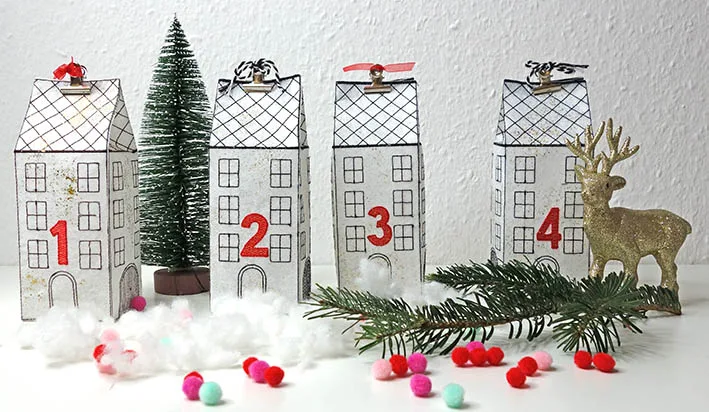 Four embroidered Advent houses with red numbers, surrounded by cotton wool, fir branches and colorful pom-poms