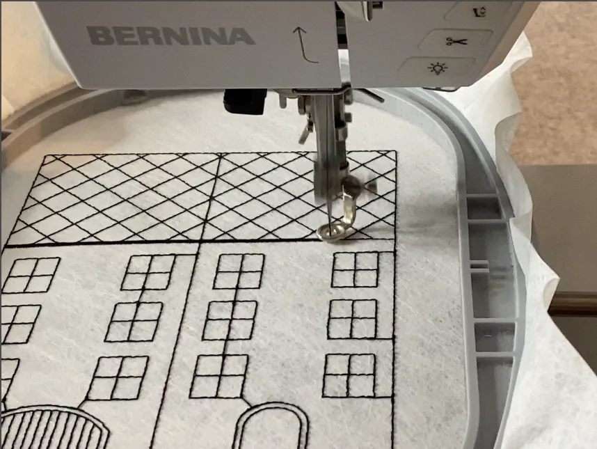 Embroidery machine stitching the facade of an ITH house with windows and roof shingles onto spunbond stabilizer